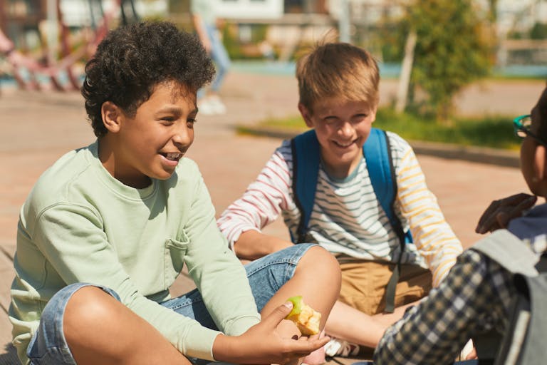 Children sitting outdoors, enjoying a fun lunch break with friends in the summer sun.